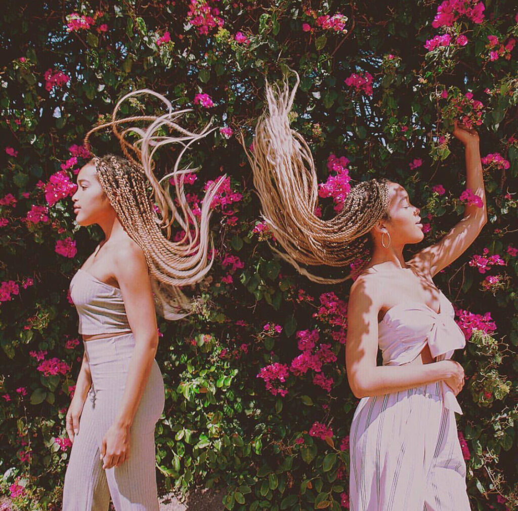 RaeCola photographed in light pink outfits in front of pink flowers