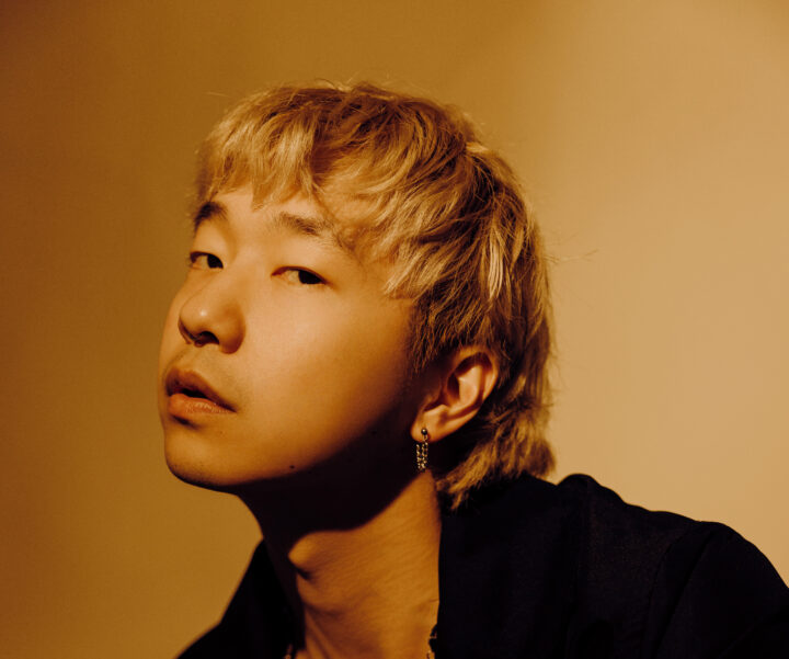 Photo of a blonde asian man with a suit jacket and no shirt underneath in a yellow light.