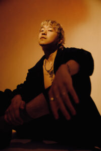 Photo of a blonde asian man with a suit jacket and no shirt underneath in a yellow light.