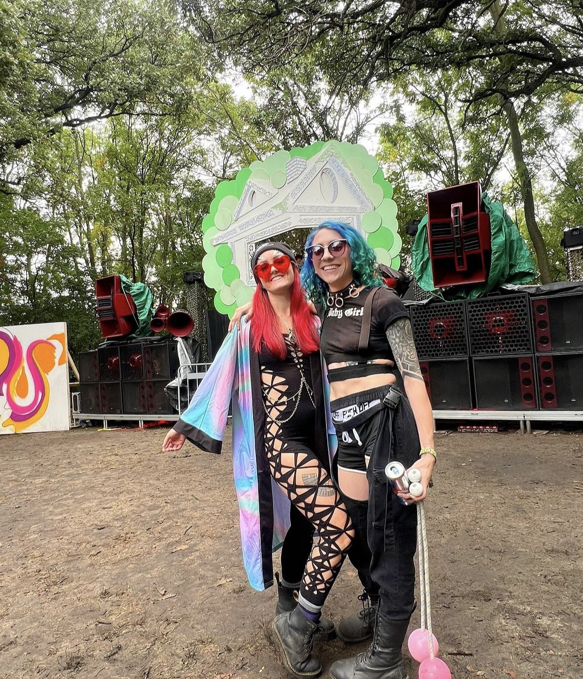 Two women, one with red hair and one with blue hari, posing for a photo in front of large speakers in the woods