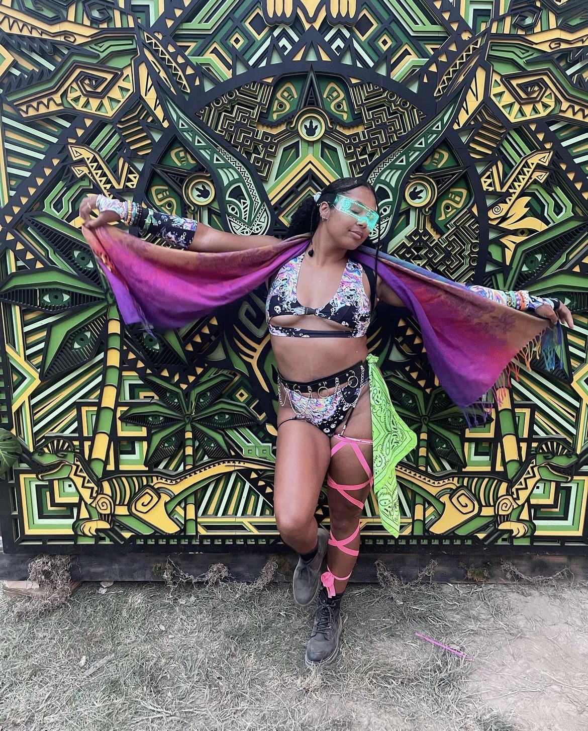 Woman wearing psychedelic two piece outfit with dangling chains and pink leg wrap holding a pashmina and wearing green glasses in front of green and yellow psychedelic art wall