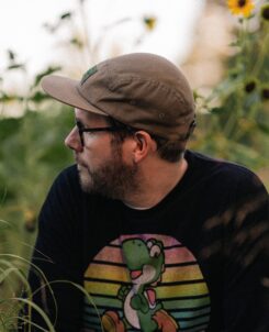 Late Night Radio headshot. Man with hat staring off camera in front of garden, wearing a hat.