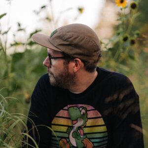 Late Night Radio headshot. Man with hat staring off camera in front of garden, wearing a hat.