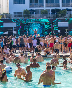 Pool party featuring "Green Velvet" DJ playing poolside while people throw inflatable beach balls in the pool.