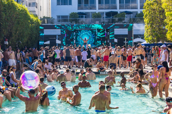 Pool party featuring "Green Velvet" DJ playing poolside while people throw inflatable beach balls in the pool.