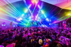Crowd of people dancing at a music festival under colorful lights.