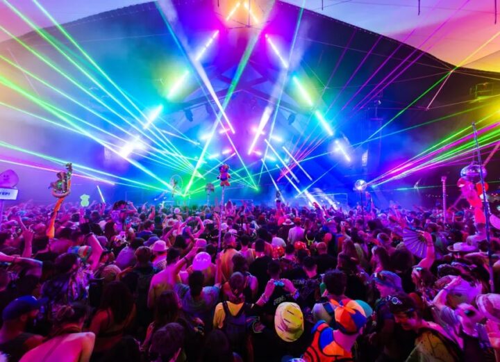 Crowd of people dancing at a music festival under colorful lights.