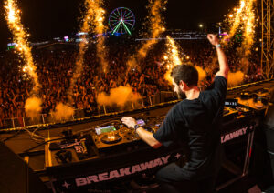 DJ on stage playing in front of a massive crowd at night, with a ferris wheel in the background