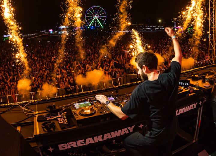 DJ on stage playing in front of a massive crowd at night, with a ferris wheel in the background