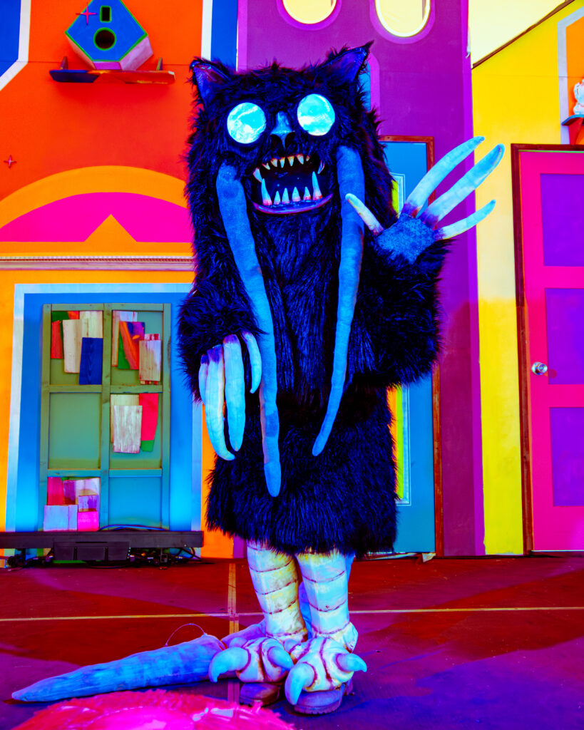 person in blue monster suit standing in front of a colorful art exhibit, waving at the camera
