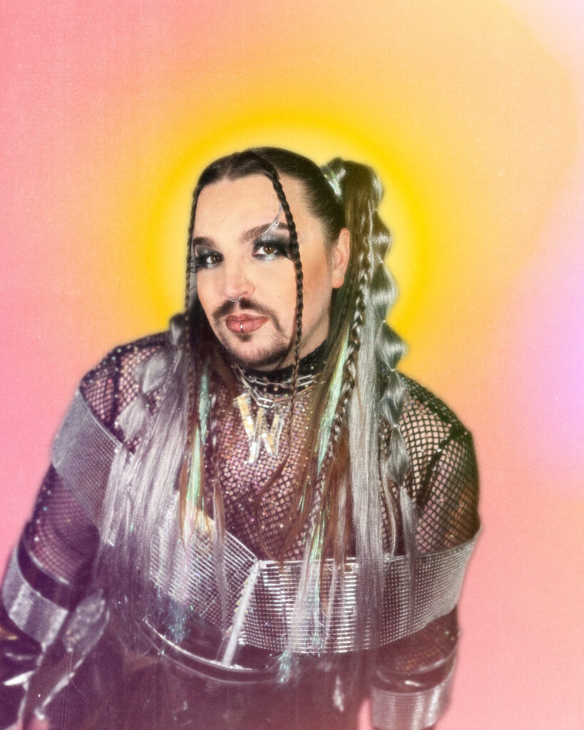 Wreckno posing in front of a pink and orange backdrop, wearing colorful makeup and pony tails.