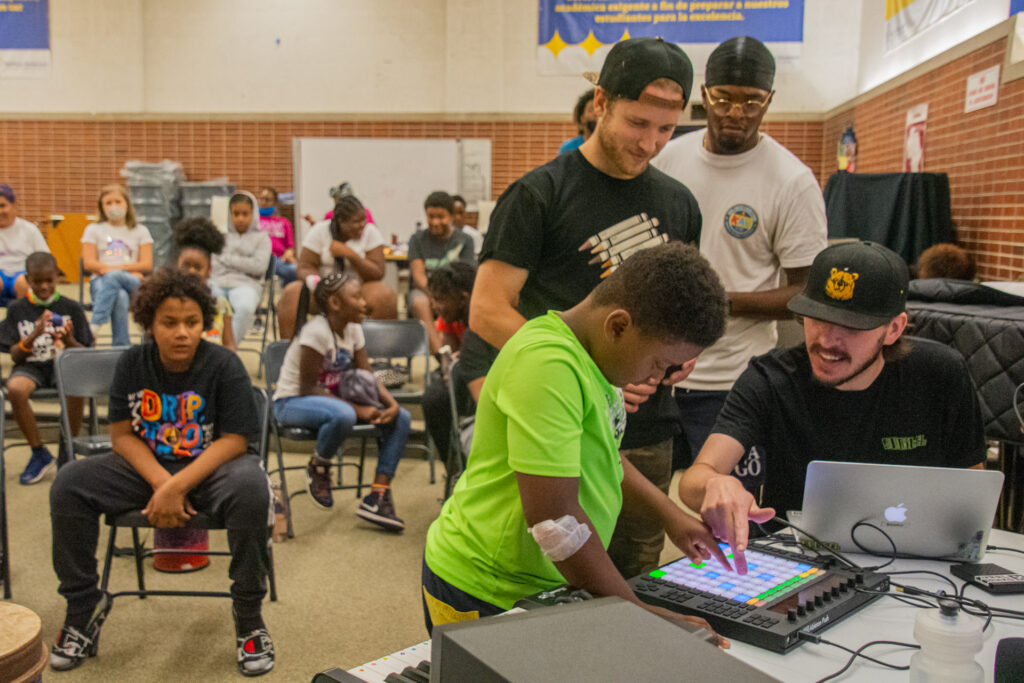 BoredomFighters hosting a music production workshop in a school classroom, teaching a kid to play with a drum pad