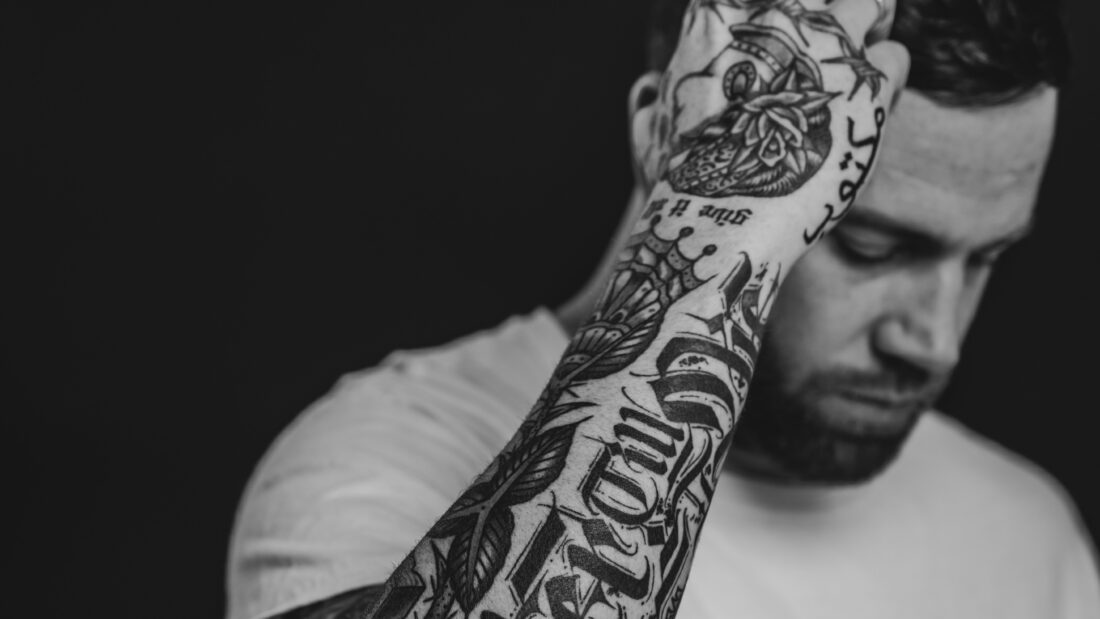 Jantsen posing in front of a black background, showing off his arm tattoos. Black and white.