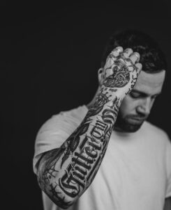 Jantsen posing in front of a black background, showing off his arm tattoos. Black and white.