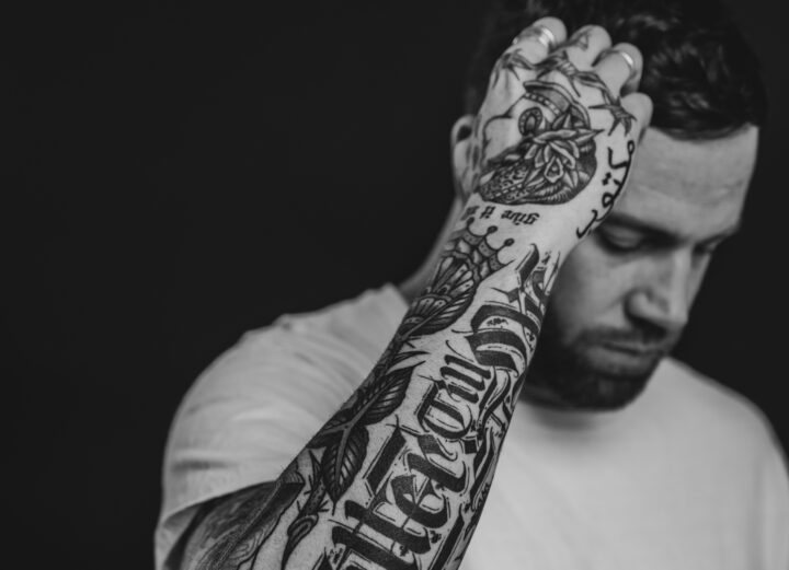 Jantsen posing in front of a black background, showing off his arm tattoos. Black and white.